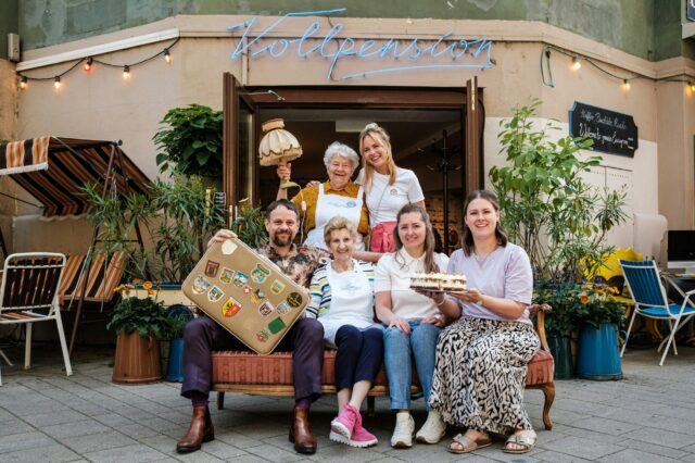 (c) Vollpension Sechs Personen vor einem Café in Wien, mit Kuchen und Koffer in der Hand.