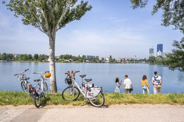 Vier Menschen stehen an einem der Badeplätze in Wien, ihre Fahrräde sind an einen Baum angelehnt.