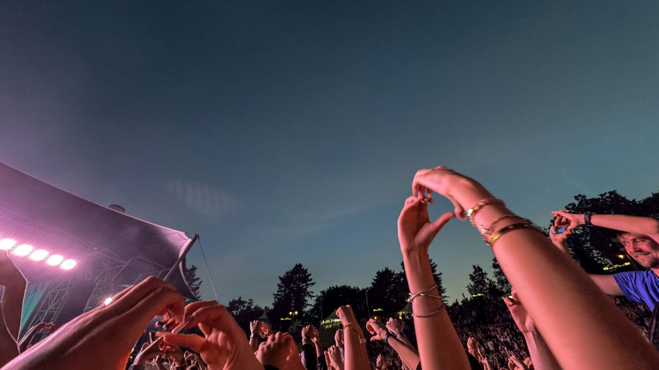 (c) Sonja Koller | 1000things Ausgelassene Festivalstimmung beim Berlin Festival Sommer 2025: Zahlreiche Menschen stehen vor einer Bühne unter freiem Himmel in der Abenddämmerung, halten Hände in die Luft und formen Herzzeichen. Die Bühne ist in pink-violettes Licht getaucht, umgeben von Bäumen und sommerlicher Atmosphäre.