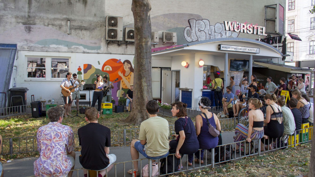 (c) Rumwurschteln Eine Gruppe Menschen vor und um einen Würstelstand in Wien herum, die einem Konzert lauschen.