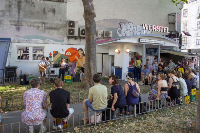 Eine Gruppe Menschen vor und um einen Würstelstand in Wien herum, die einem Konzert lauschen.