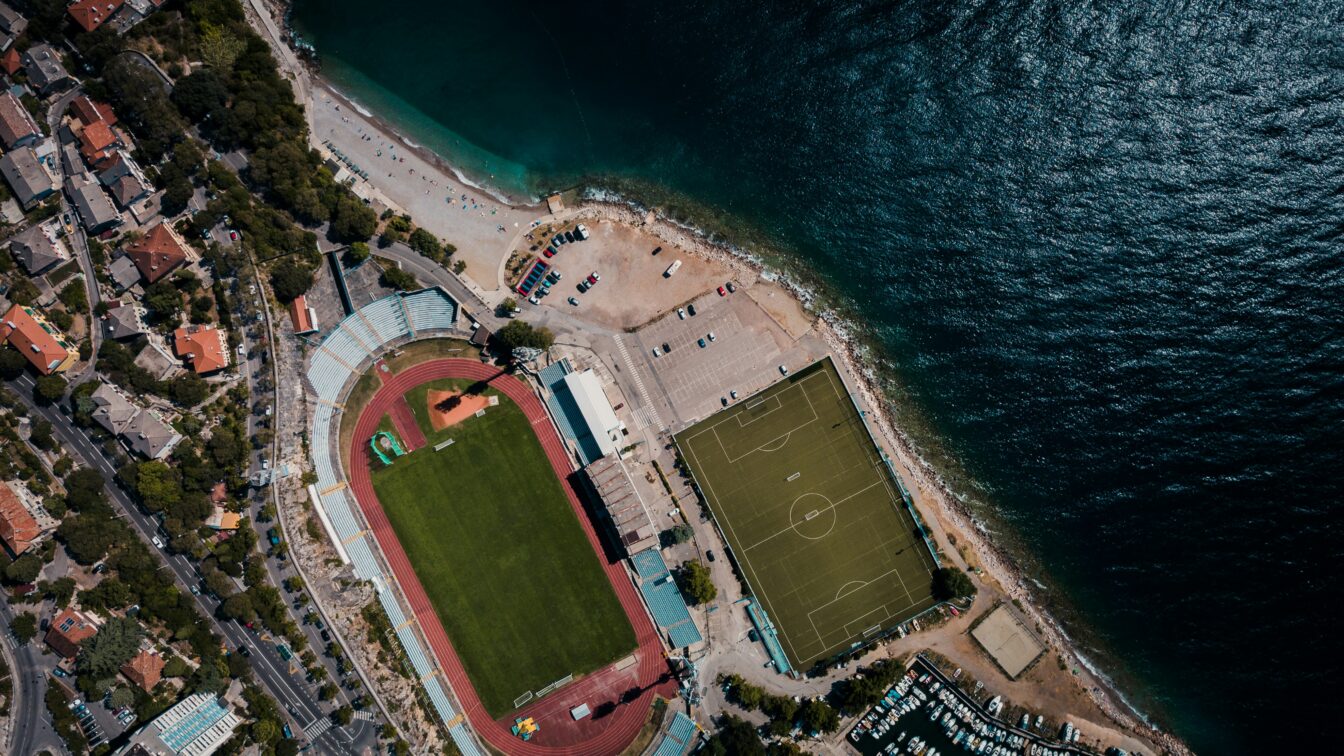 (c) Daniel James | Unsplash Eine Stadt am Meer mit Fußballstadion und Sportplatz aus der Vogelperspektive.