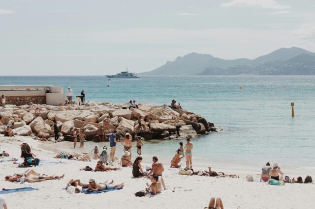 Menschen genießen einen sonnigen Tag an einem belebten Sandstrand in Südfrankreich. Einige liegen auf Handtüchern, andere baden im klaren türkisblauen Meer oder sitzen auf großen Felsen. Im Hintergrund ist ein luxuriöses weißes Schiff auf dem Wasser zu sehen, dahinter eine hügelige Küstenlandschaft. Die Atmosphäre wirkt entspannt und sommerlich.