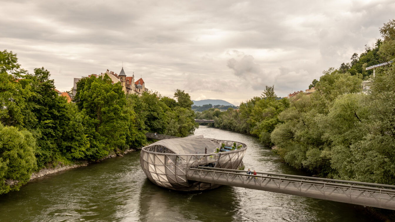 (c) Sonja Koller | 1000things Blick auf die Murinsel in Graz an einem bewölkten Sommertag – modernes, schalenförmiges Bauwerk auf der Mur, umgeben von grünen Bäumen, historischer Architektur im Hintergrund und Spaziergänger*innen auf der Brücke