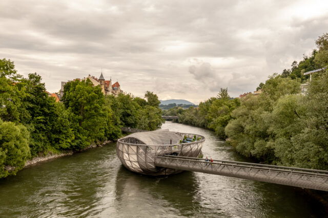 Blick auf die Murinsel in Graz an einem bewölkten Sommertag – modernes, schalenförmiges Bauwerk auf der Mur, umgeben von grünen Bäumen, historischer Architektur im Hintergrund und Spaziergänger*innen auf der Brücke