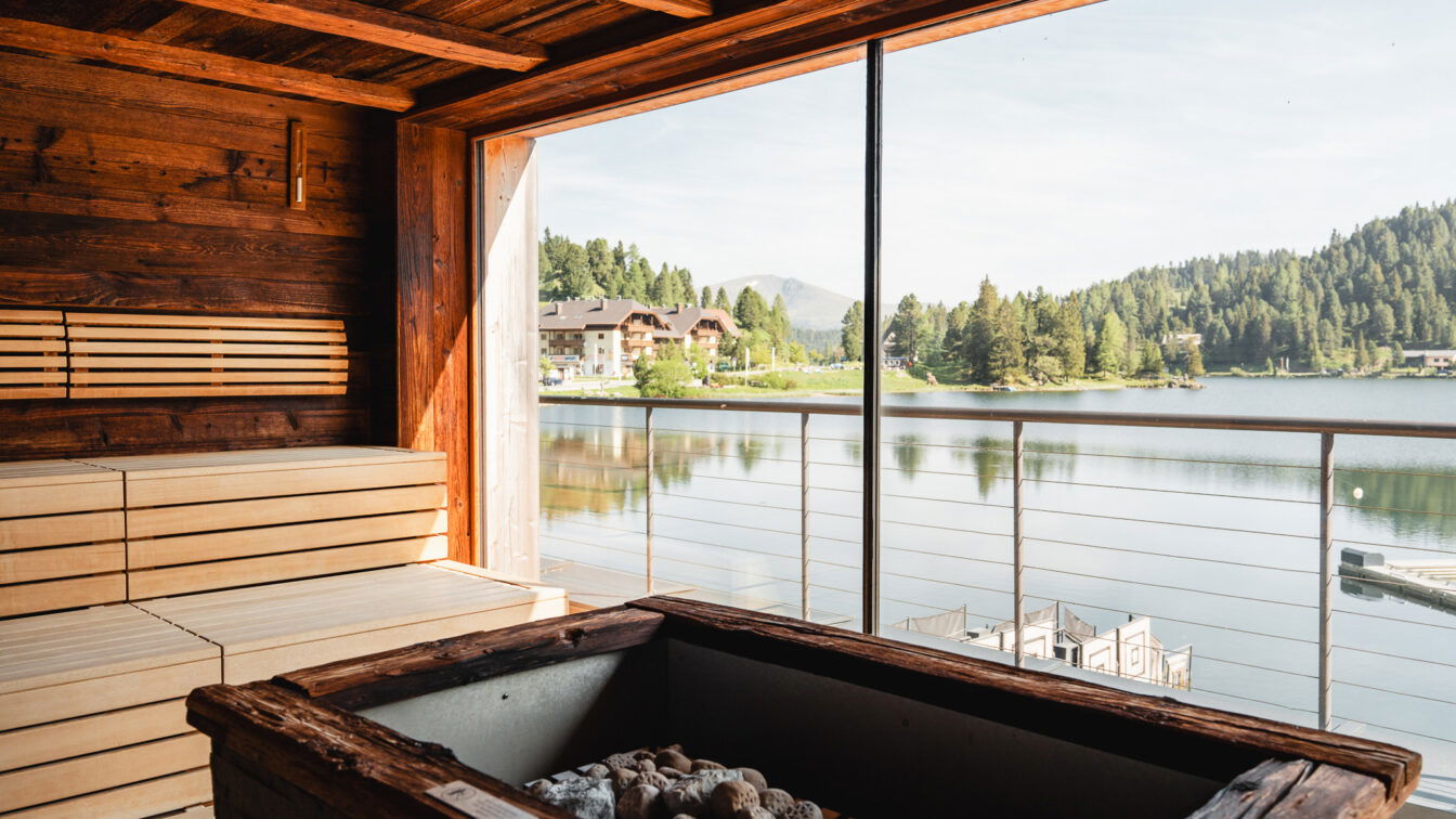 (c) Lilli Wermuth | 1000things Blick aus einer Sauna heraus mit Glas-Wand und Sicht auf einen See mit Bergen im Hintergrund.