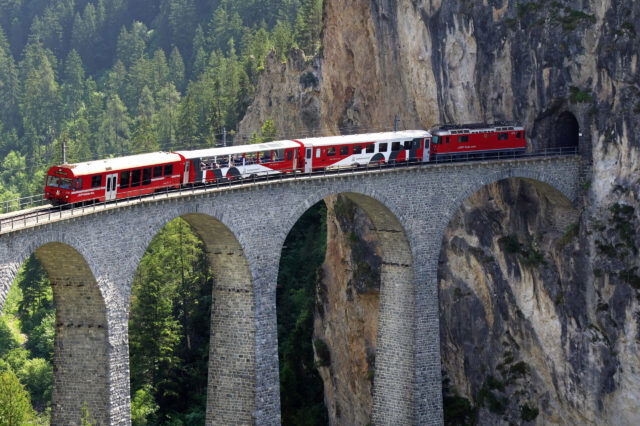 Ein rot-weißer Zug, der vorbei an einer grünen Landschaft über ein Landwasserviadukt fährt.