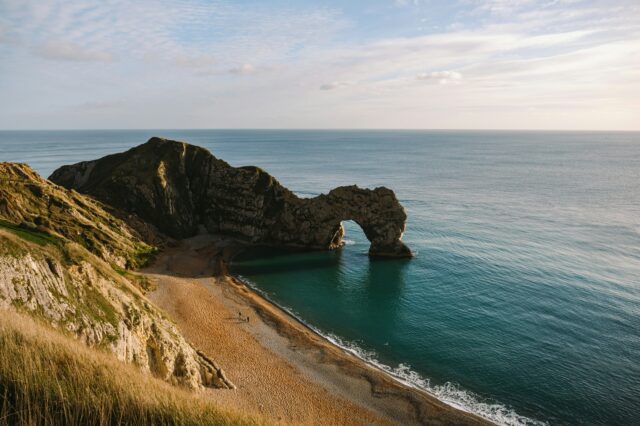 Durdle Door in Wareham, Großbritannien. (c) Pauline Herpel | Unsplash Blick von einer Klippe aus auf eine Felsenformatione, die ins Wasser ragt.