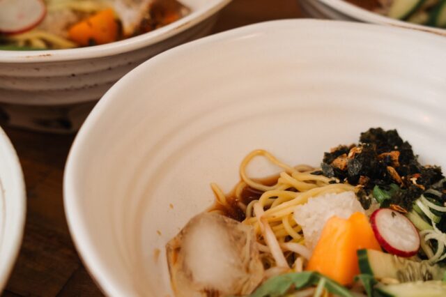 (c) Julia Riedesser | 1000things Drei weiße Keramikschalen mit modernen Ramen-Gerichten: feine Weizennudeln in aromatischer Brühe, garniert mit knusprigem Seetang, Radieschenscheiben, Frühlingszwiebeln und buntem Gemüse – zeitgemäße japanische Küche in stilvollem Ambiente.