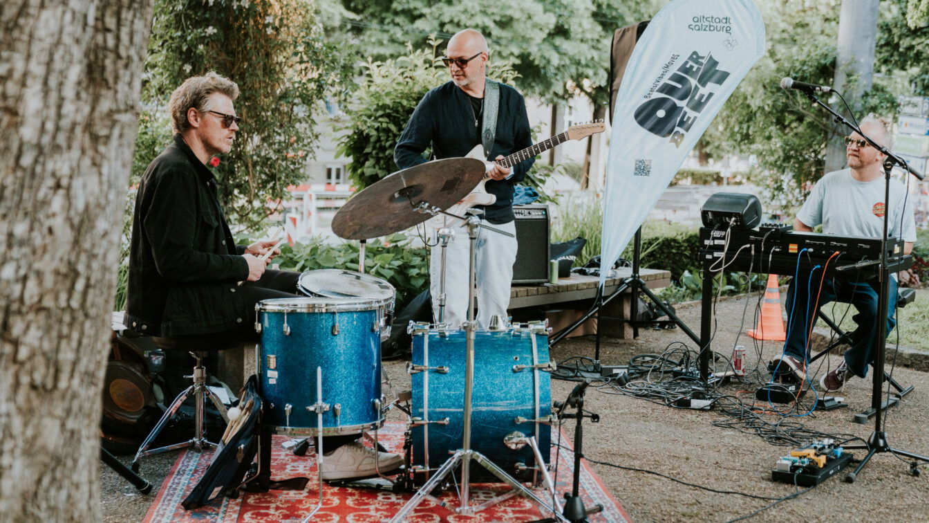 (c) fraeuleinflora.at Live-Musik im Schanzlpark Salzburg – Band mit Schlagzeug, Gitarre und Keyboard bei Open-Air-Konzert der Veranstaltungsreihe 'Altstadt Salzburg Über d’Gass', stimmungsvolle Atmosphäre unter freiem Himmel.