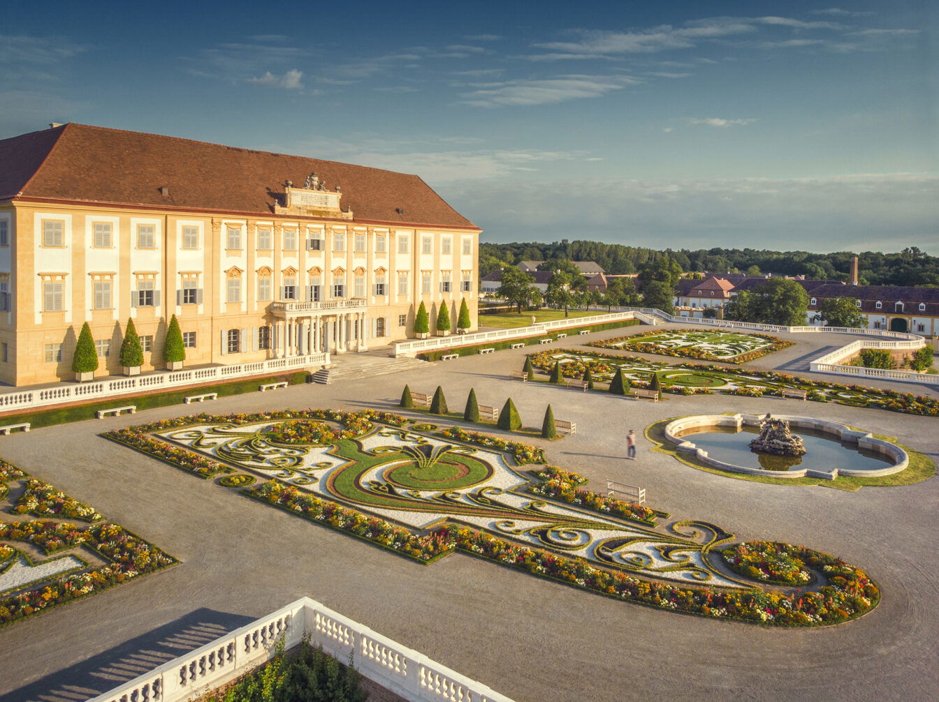 Blick auf das barocke Schloss Hof mit seiner prachtvollen Fassade und dem kunstvoll angelegten Schlossgarten. Der symmetrische Garten ist mit blühenden Blumenbeeten, Hecken, Skulpturen und einem Springbrunnen gestaltet. Die Szene ist in warmes Sonnenlicht getaucht und vermittelt eine Atmosphäre historischer Eleganz und Ruhe.