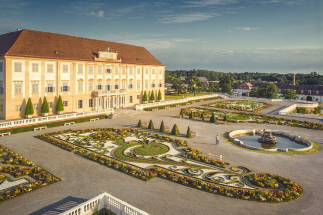 Blick auf das barocke Schloss Hof mit seiner prachtvollen Fassade und dem kunstvoll angelegten Schlossgarten. Der symmetrische Garten ist mit blühenden Blumenbeeten, Hecken, Skulpturen und einem Springbrunnen gestaltet. Die Szene ist in warmes Sonnenlicht getaucht und vermittelt eine Atmosphäre historischer Eleganz und Ruhe.