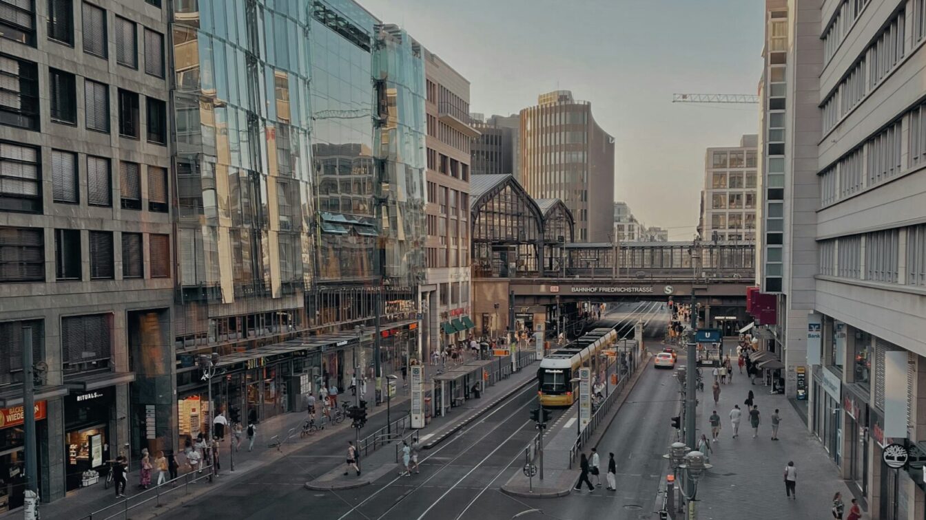 (c) Shobhit Sharma | Unsplash Foto einer Straßenschlucht, im Hintergrund sieht man Bahnschienen mit der Aufschrift Bahnhof Friedrichstraße.