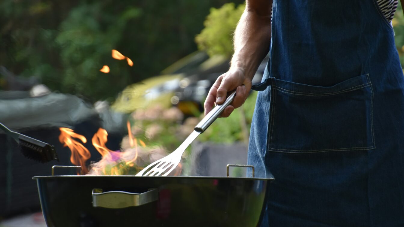 (c) Vincent Keiman | Unsplash Eine Person mit Grillschürze und Wender steht an einem Feuer-Griller