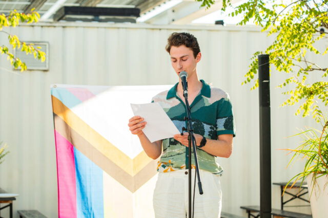 (c) Neuwirth Person mit buntem Hemd auf einer flachen Bühne, die einen Text liest, mit Pride-Flagge im Hintergrund.