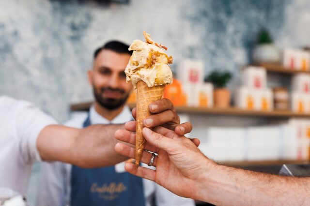 (c) Adrian Almasan Eine Hand, die eine Kugel Eis in der Waffel überreicht mit einem Mann im Hintergrund.