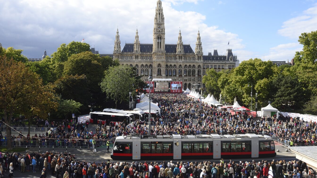 (c) Johannes Zinner | Wiener Linien Große Menschenmenge bei der Tram-WM vor dem Wiener Rathaus, zahlreiche Besucher versammeln sich rund um eine moderne Straßenbahn und mehrere Infostände, im Hintergrund das historische Rathausgebäude mit seinen Türmen