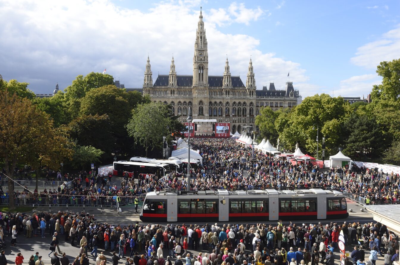 Große Menschenmenge bei der Tram-WM vor dem Wiener Rathaus, zahlreiche Besucher versammeln sich rund um eine moderne Straßenbahn und mehrere Infostände, im Hintergrund das historische Rathausgebäude mit seinen Türmen