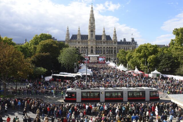 Große Menschenmenge bei der Tram-WM vor dem Wiener Rathaus, zahlreiche Besucher versammeln sich rund um eine moderne Straßenbahn und mehrere Infostände, im Hintergrund das historische Rathausgebäude mit seinen Türmen