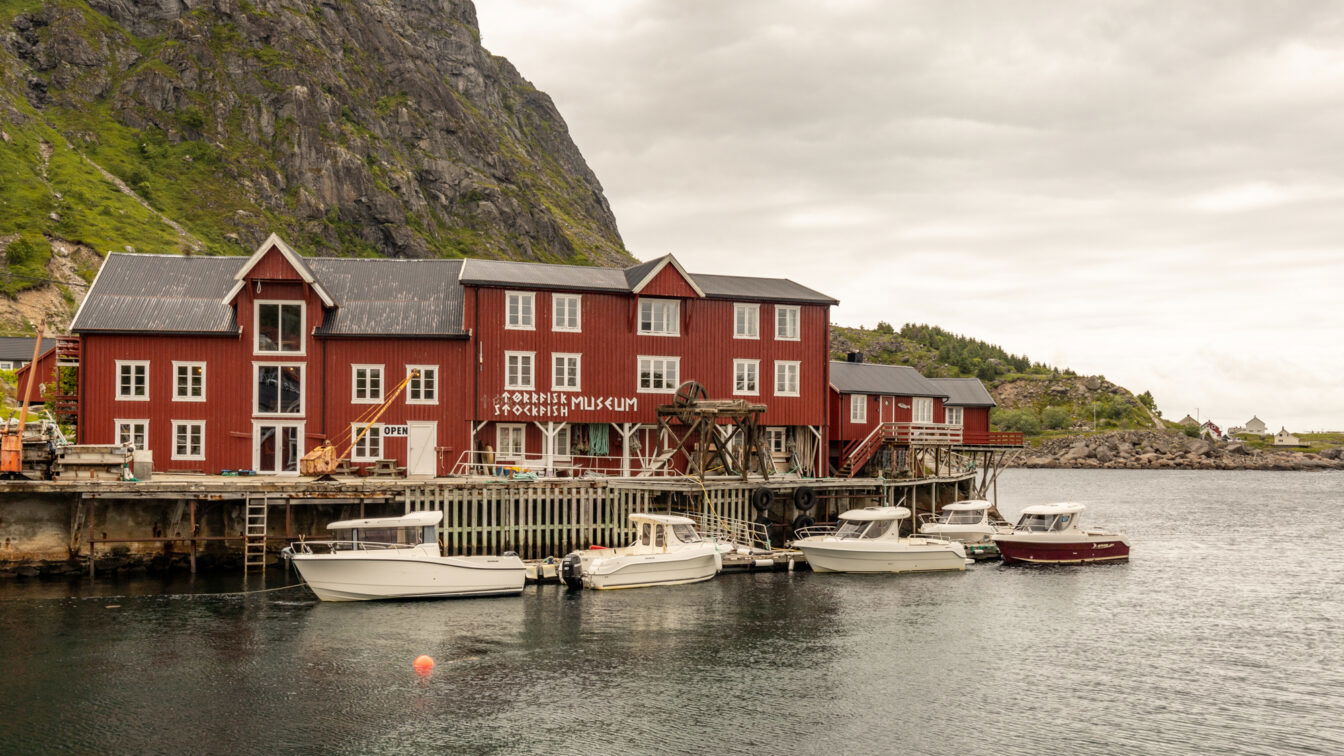 (c) Sonja Koller | 1000things Å i Lofoten, Norwegen – historische rote Holzhäuser am Wasser mit dem Norwegischen Stockfischmuseum, umgeben von steilen Felswänden und Booten im ruhigen Hafen bei bewölktem Himmel.