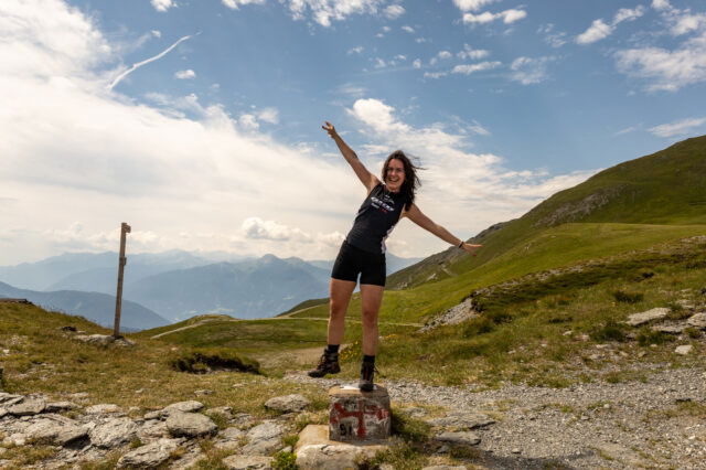 Person in Sportkleidung steht lachend auf einem Grenzstein am Pass während der Alpenüberquerung, umgeben von grünen Hügeln und einem weiten Bergpanorama unter blauem Himmel mit leichten Wolken.