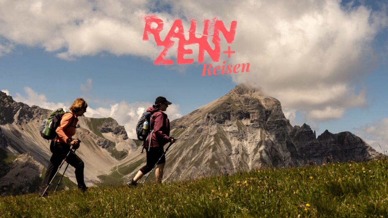 (c) Sonja Koller | 1000things Zwei wandernde Personen mit Rucksäcken und Wanderstöcken überqueren eine grüne Wiese vor einer beeindruckenden Bergkulisse mit steilen Felswänden und Wolken am Himmel. Im oberen Bildbereich steht in roter Schrift „Rauzen + Reisen“.