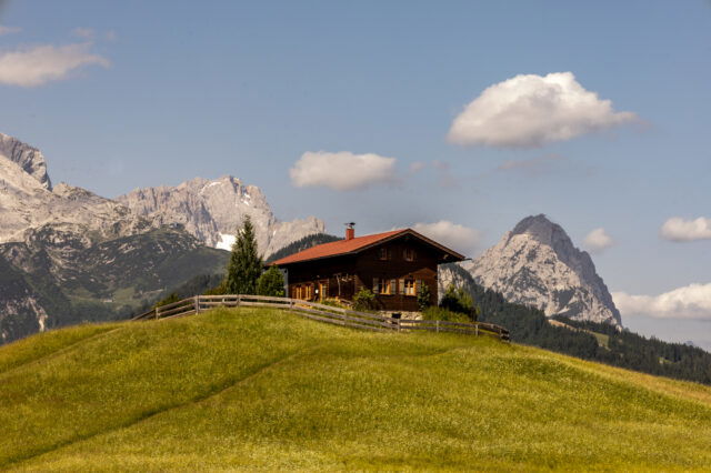 Holzhaus auf einer grünen Almwiese bei Garmisch-Partenkirchen, im Hintergrund schroffe Alpen mit einzelnen Schneefeldern und blauer Himmel mit Wolken, idealer Startpunkt für eine Alpenüberquerung nach Mittenwald.