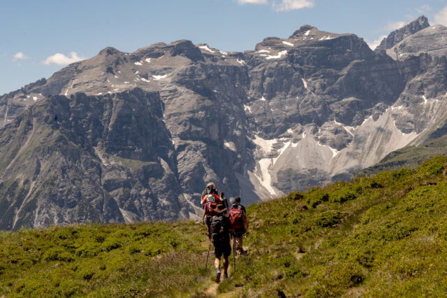 (c) Sonja Koller | 1000things Wandergruppe mit Rucksäcken und Stöcken auf einem schmalen Almweg, der auf ein mächtiges, graues Felsmassiv mit Schneefeldern und blauem Himmel in den Tiroler Alpen zuführt.