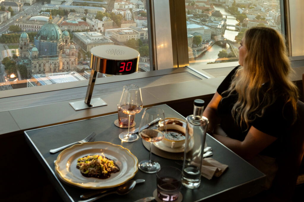 Romantisches Dinner mit Panoramablick über Berlin im Fernsehturm-Restaurant – perfekte Date-Idee in Berlin. Eine Frau genießt bei Sonnenuntergang den Ausblick auf den Berliner Dom und die Spree, während auf dem Tisch edle Speisen, Wein und stilvolles Ambiente zum Verweilen einladen