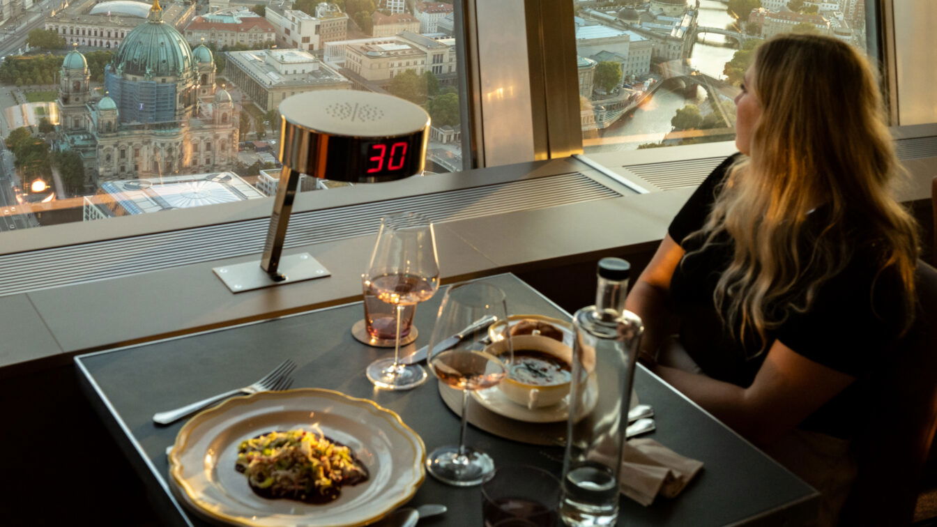 Romantisches Dinner mit Panoramablick über Berlin im Fernsehturm-Restaurant – perfekte Date-Idee in Berlin. Eine Frau genießt bei Sonnenuntergang den Ausblick auf den Berliner Dom und die Spree, während auf dem Tisch edle Speisen, Wein und stilvolles Ambiente zum Verweilen einladen