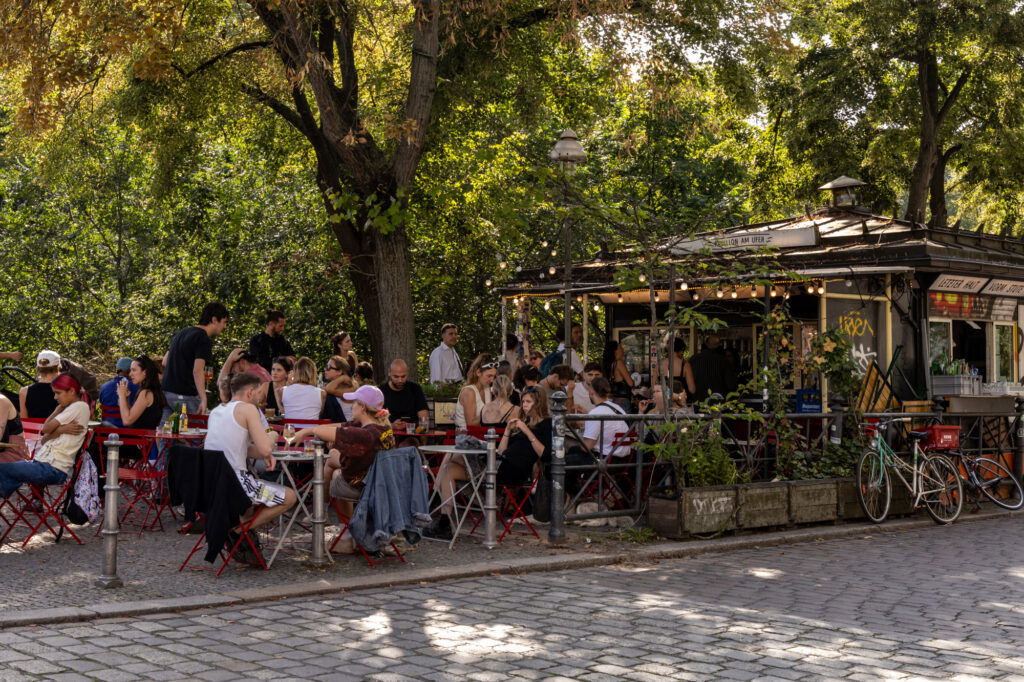Lebendiges Berliner Sommer-Event im August mit vielen Menschen, die an roten Bistrotischen vor einem charmanten Kiosk namens ‚Salon am Ufer‘ sitzen. Unter schattigen Bäumen und Lichterketten genießen Gäste Snacks und Getränke – typisches Kiezleben bei Events in Berlin im August.