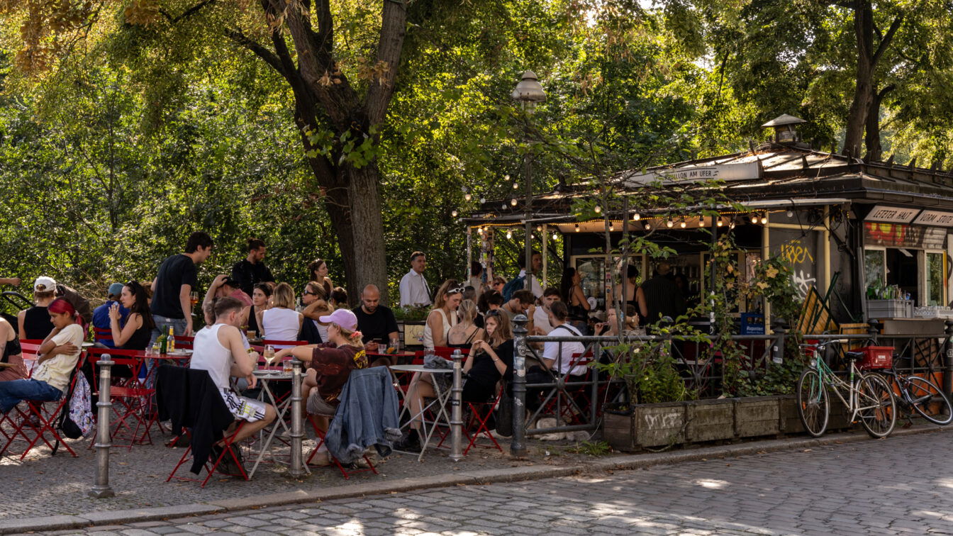 (c) Sonja Koller | 1000things Lebendiges Berliner Sommer-Event im August mit vielen Menschen, die an roten Bistrotischen vor einem charmanten Kiosk namens ‚Salon am Ufer‘ sitzen. Unter schattigen Bäumen und Lichterketten genießen Gäste Snacks und Getränke – typisches Kiezleben bei Events in Berlin im August.