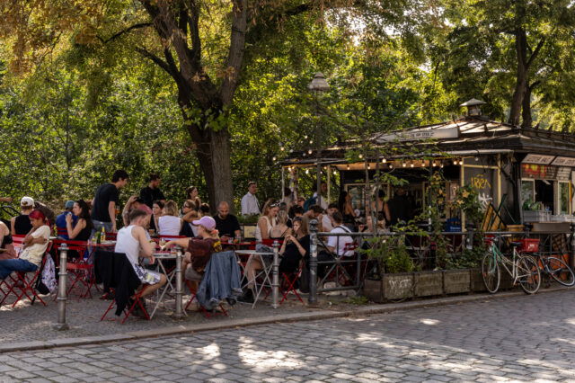 Lebendiges Berliner Sommer-Event im August mit vielen Menschen, die an roten Bistrotischen vor einem charmanten Kiosk namens ‚Salon am Ufer‘ sitzen. Unter schattigen Bäumen und Lichterketten genießen Gäste Snacks und Getränke – typisches Kiezleben bei Events in Berlin im August.