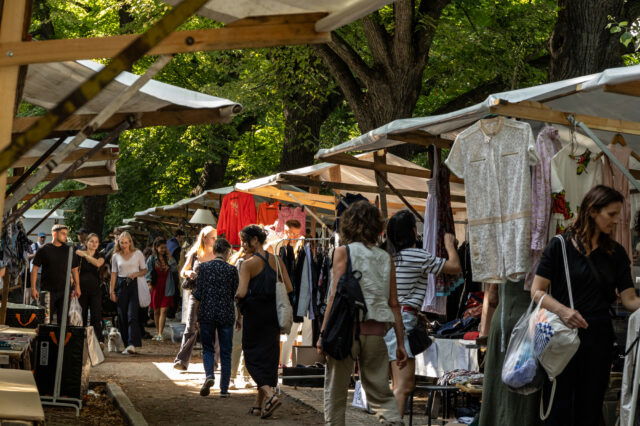 Belebter Second-Hand-Markt in Berlin unter großen schattenspendenden Bäumen. Zahlreiche Menschen schlendern entlang der Stände mit Kleidung, die an Holzgestellen hängt. Die Atmosphäre wirkt entspannt und sommerlich, mit vielfältiger Mode und neugierigen Besucher*innen.