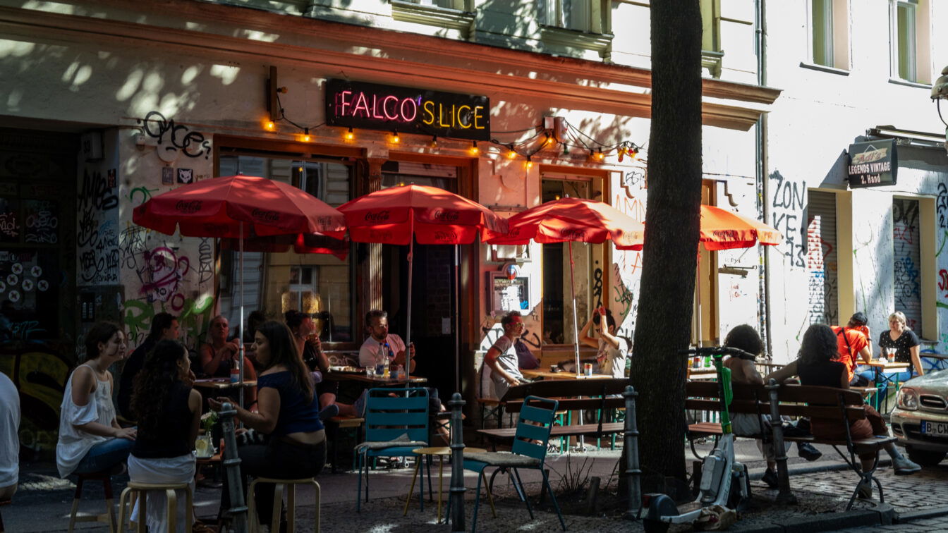 (c) Sonja Koller | 1000things Sommerszene vor der Pizzeria ‚Falco’s Slice‘ in Berlin: Menschen sitzen entspannt unter roten Sonnenschirmen auf dem Gehweg, umgeben von Graffiti, bunten Stühlen und urbanem Flair – ein typischer Moment für einen Berlin Urlaub abseits der Touristenpfade.