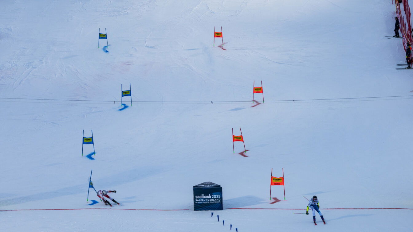 Zwei alpine Skirennläufer:innen durchfahren parallel einen Riesenslalom-Parcours während eines Qualifikationsrennens oder Wettbewerbs. Im Zielbereich steht ein Schild mit der Aufschrift „Saalbach 2025 SalzburgerLand“, dem Austragungsort der FIS Alpinen Ski-Weltmeisterschaften 2025, die auch als Vorbereitung auf die Olympischen Winterspiele 2026 in Mailand-Cortina dienen.