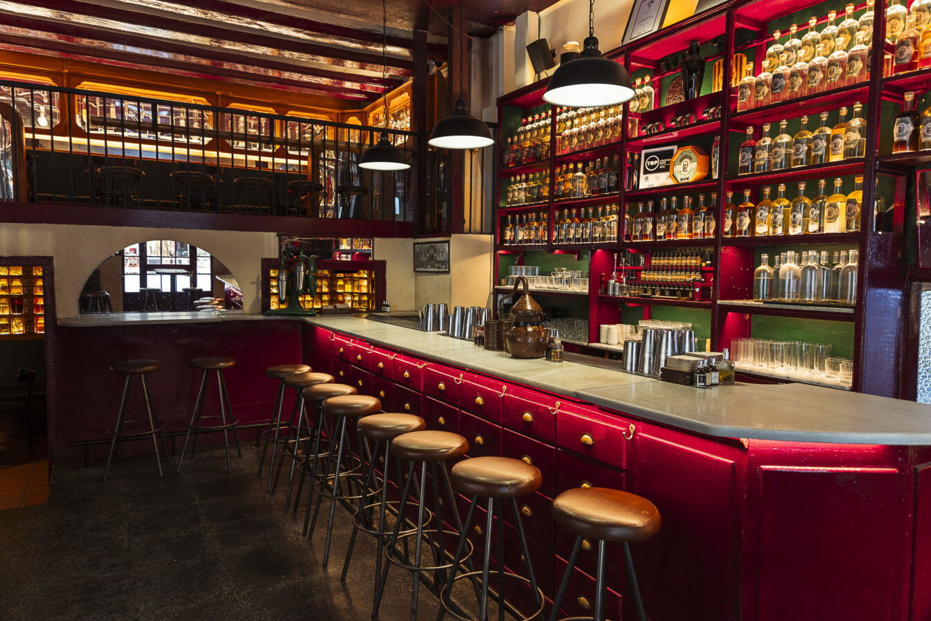 Eine Bar mit beleuchteten Regalen und vielen Flaschen, rotem Thresen und Barhockern aus Leder.