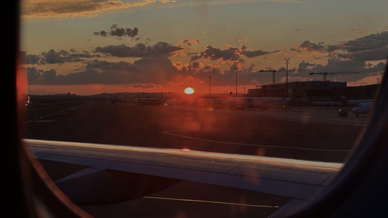 (c) Sonja Koller | 1000things Blick aus dem Fenster eines Flugzeugs beim Sonnenuntergang auf dem Rollfeld eines Flughafens. Die Szene zeigt parkende Flugzeuge, den Flughafenbetrieb mit Gebäuden und Kränen im Hintergrund. Das Bild symbolisiert eine Wartezeit oder mögliche Flugverspätung – ein typischer Anlass, bei dem Passagieren Fluggastrechte zustehen können, etwa Entschädigungen bei verspäteten oder gestrichenen Flügen.