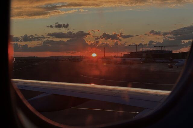 Blick aus dem Fenster eines Flugzeugs beim Sonnenuntergang auf dem Rollfeld eines Flughafens. Die Szene zeigt parkende Flugzeuge, den Flughafenbetrieb mit Gebäuden und Kränen im Hintergrund. Das Bild symbolisiert eine Wartezeit oder mögliche Flugverspätung – ein typischer Anlass, bei dem Passagieren Fluggastrechte zustehen können, etwa Entschädigungen bei verspäteten oder gestrichenen Flügen.