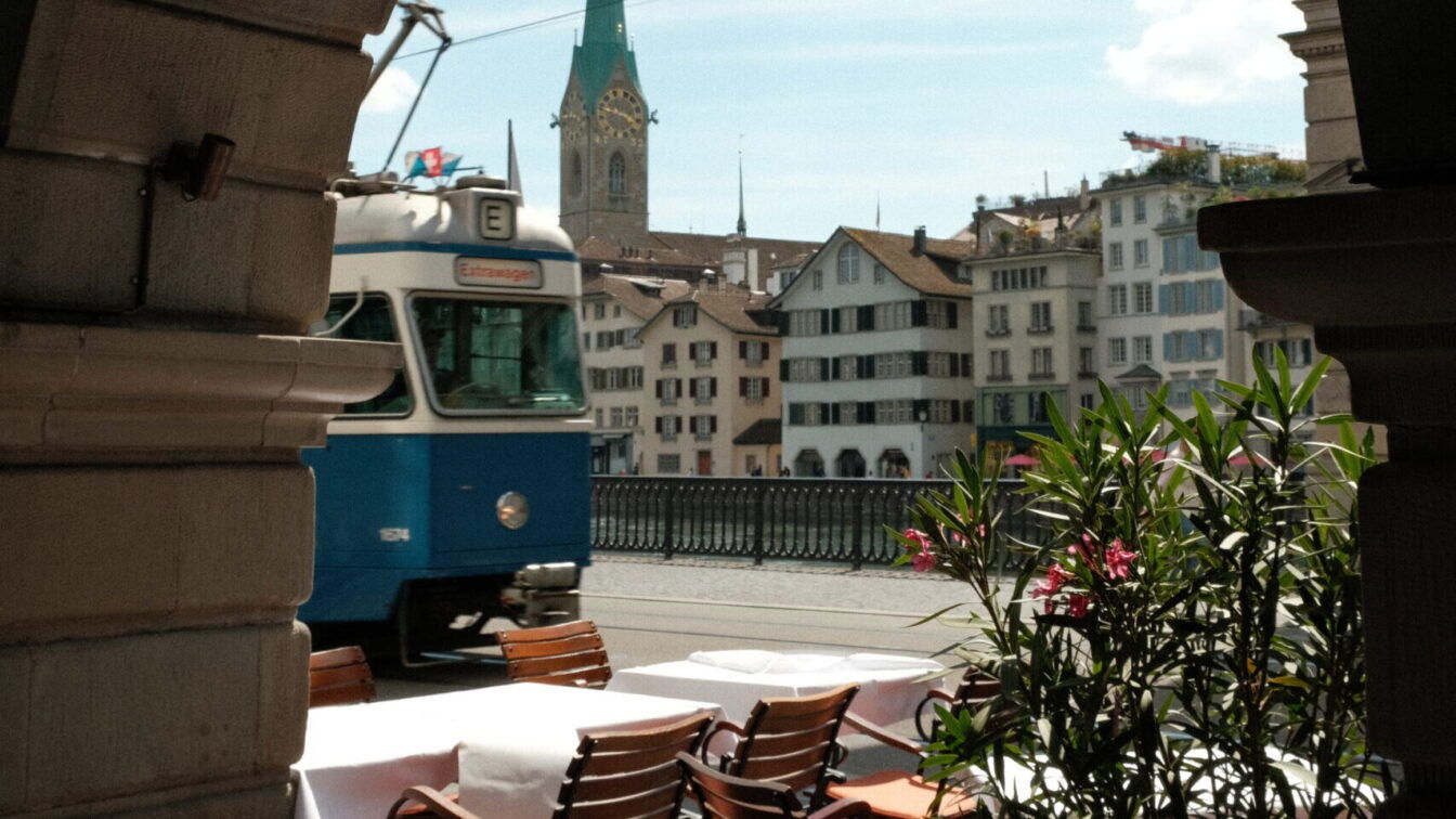 (c) Barbara Palcher | 1000things Blick durch einen steinernen Torbogen in Zürich auf einen blauen Tram, dahinter die Fraumünsterkirche mit ihrem markanten grünen Turm. Im Vordergrund stehen gedeckte Restauranttische mit Stühlen auf einer Terrasse am Fluss.