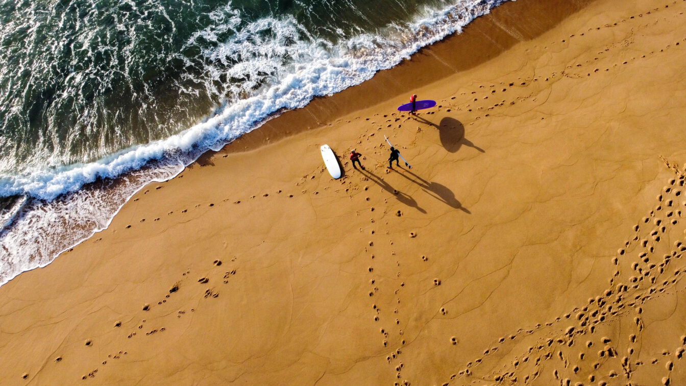 (c) GoBeyond Vogelperspektive auf drei surfende Personen am Strand vor dem Wasser in Portugal