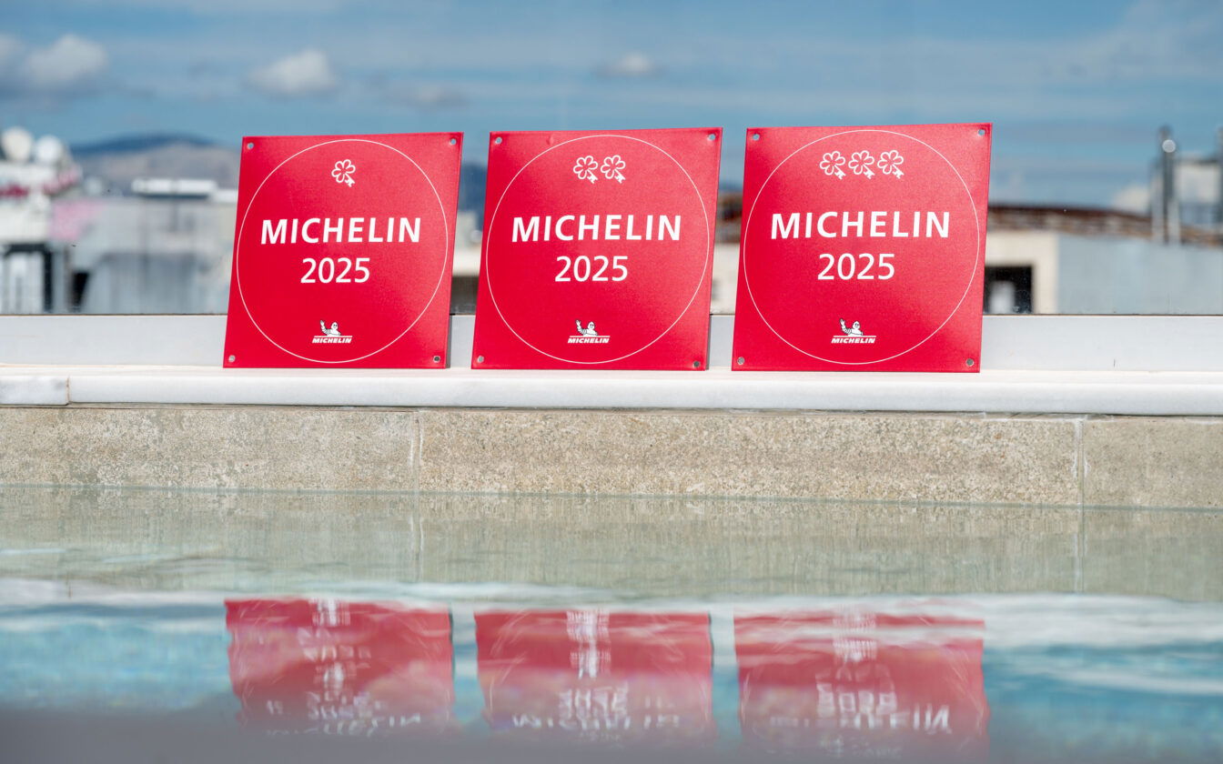 Drei rote MICHEIN 2025-Schilder mit einem, zwei und drei stilisierten Blumen-Symbolen (für Sterne) stehen nebeneinander am Rand eines Pools. Die Schilder spiegeln sich im Wasser. Im Hintergrund ist ein unscharfer Stadt- oder Dachlandschaftsausblick mit blauem Himmel zu sehen.