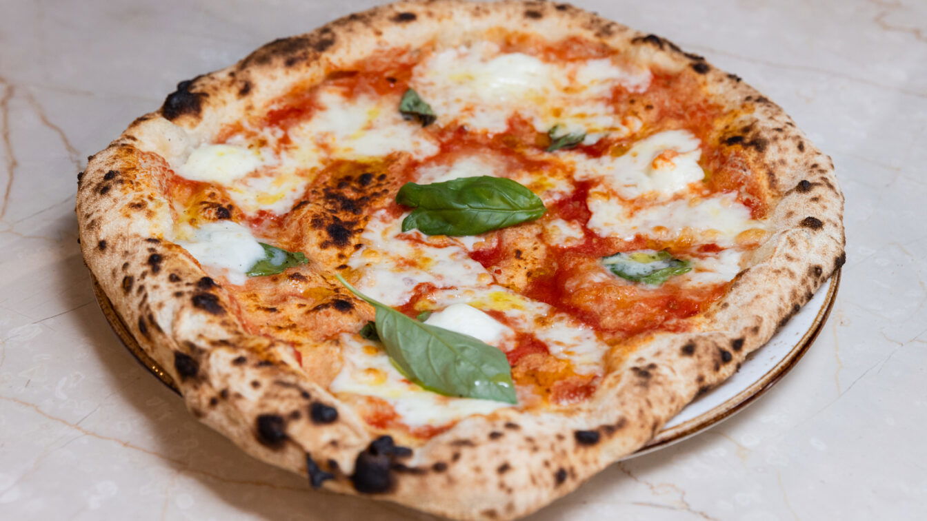 (c) ROBIN CONSULT Fellner Nahaufnahme einer frisch gebackenen Pizza Margherita mit geschmolzenem Mozzarella, Tomatensauce und frischen Basilikumblättern auf einem hellen Marmortisch.