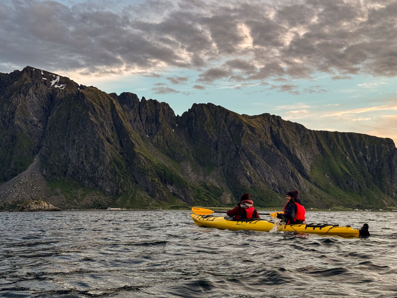 Kajaktour bei Sonnenuntergang vor dramatischer Bergkulisse auf den Lofoten, Norwegen – zwei Personen im gelben Doppelkajak paddeln durch ruhige Gewässer inmitten unberührter Natur.