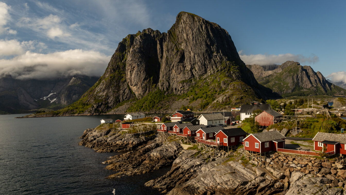 (c) Sonja Koller | 1000things Traditionelle rote Fischerhütten (Rorbuer) in malerischer Küstenlandschaft von Hamnøy auf den Lofoten, Norwegen – mit dramatischer Bergkulisse und klarer Meeresluft unter blauem Himmel.