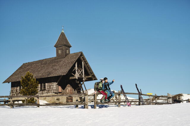 Zwei Personen sitzen auf einem Zaun und zeigen in die Ferne. Man sieht eine Winterlandschaft und hinter ihnen eine Hütte.