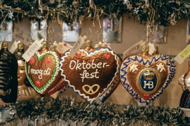 Herzförmige Lebkuchen-Ornamente hängen an Haken in einem festlich dekorierten Verkaufsstand. Die Herzen sind mit buntem Zuckerguss verziert und tragen Aufschriften wie „Oktoberfest“, „I mog di“ und das Hofbräuhaus-Logo „HB“. Preisschilder in Euro sind sichtbar. Im Hintergrund sind Tannengirlanden und weitere weihnachtliche Dekorationen zu sehen.
