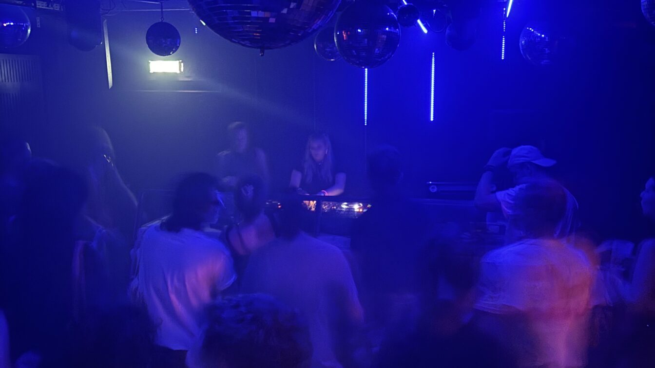 (c) Sonja Koller | 1000things Eine Szene aus einem belebten Nachtclub in Europa: Menschen tanzen unter blauem und violettem Licht, während eine DJane im Hintergrund Musik auflegt. Mehrere Diskokugeln hängen von der Decke und reflektieren das Licht, was eine lebhafte und energiegeladene Atmosphäre schafft. Die Bewegungen der Menschen wirken leicht verschwommen, was auf Tanzen und Bewegung hindeutet.
