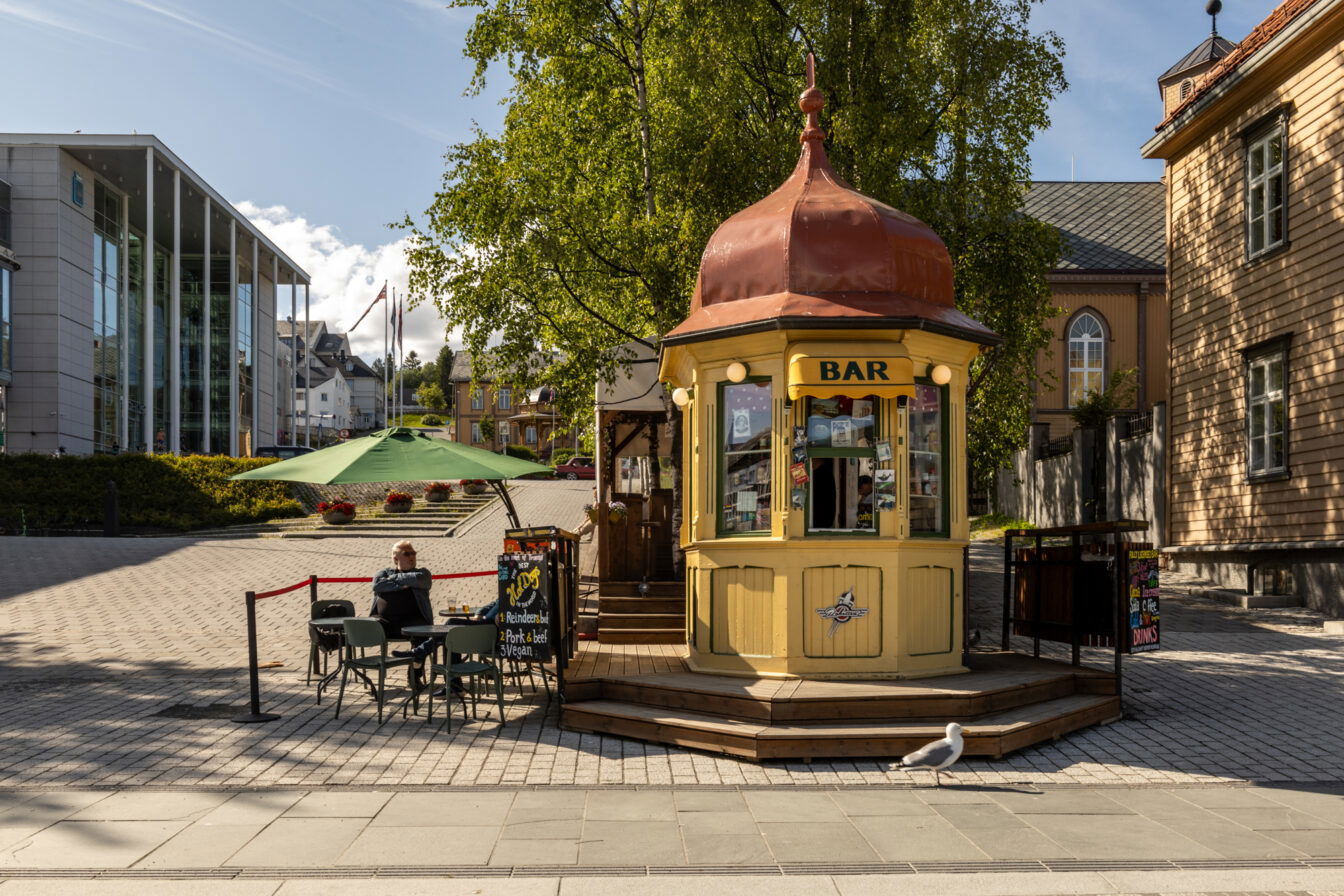 Kleiner, historischer Kiosk mit rot-gelbem Zwiebeldach in der Fußgängerzone von Tromsø, bekannt als Raketten Bar & Pølse. Eine Tafel bewirbt Hotdogs und Getränke. Davor sitzt ein Gast an einem kleinen Tisch unter einem grünen Sonnenschirm. Umgeben von klassischen und modernen Gebäuden, darunter das moderne Kulturhaus links und eine Kirche im Hintergrund. Eine Möwe spaziert im Vordergrund auf dem Bürgersteig.