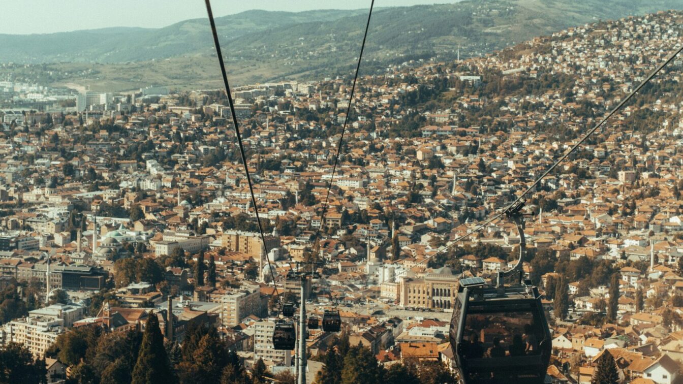 (c) Azur Golic | unsplash Blick aus einer Seilbahn auf Sarajevo mit zahlreichen Häusern, Hotels und Unterkünften, die sich über die Stadt und die umliegenden Hügel erstrecken.